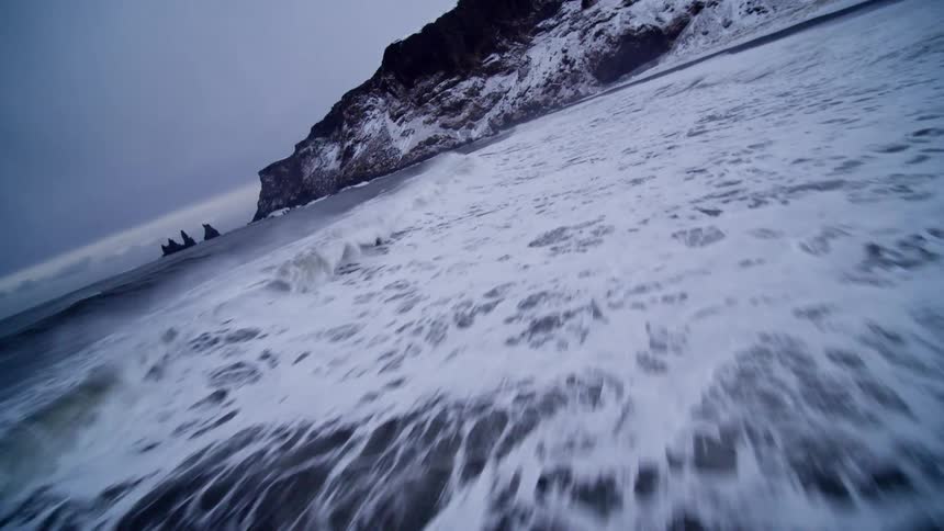 A powerful ocean wave washes over the black sand beach in an immersive slow-motion shot