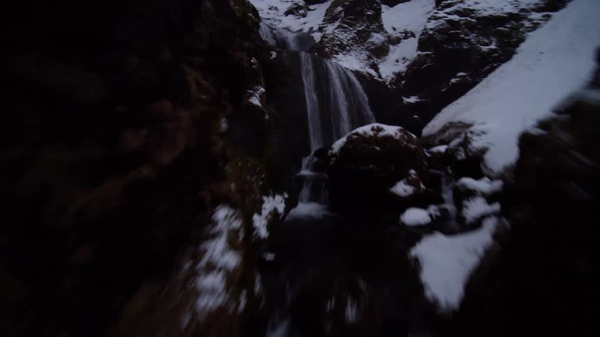A fast-paced FPV drone flight follows a dark river upstream through a snowy canyon, ascending a series of dramatic waterfalls