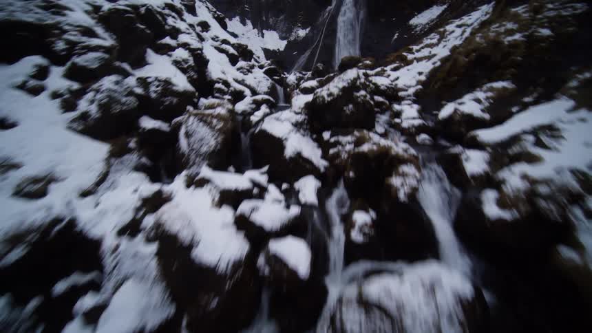 A thrilling first-person view drone flight follows a winding river upstream through a snow-covered canyon, culminating at a powerful waterfall