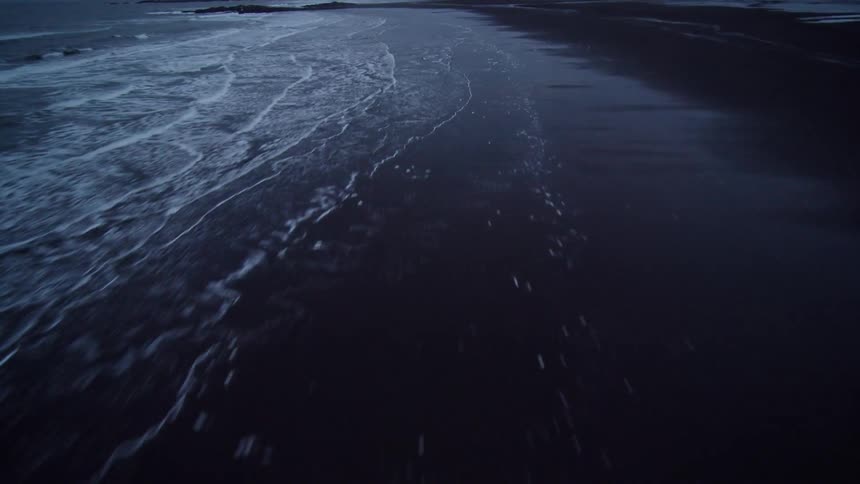 An exhilarating aerial flight over a snowy mountain ridge leads to a dramatic, icy black sand beach