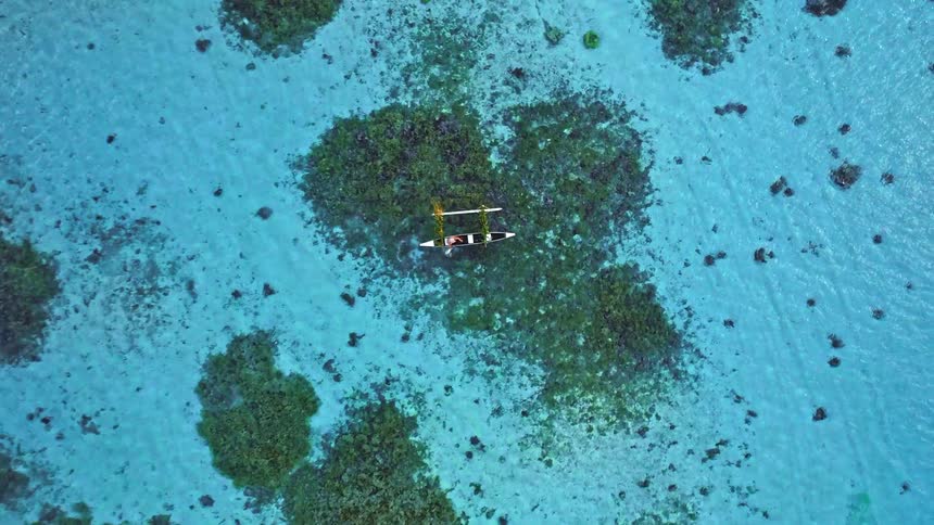 An aerial top-down view of a traditional outrigger canoe floating in a crystal-clear turquoise lagoon over a coral reef