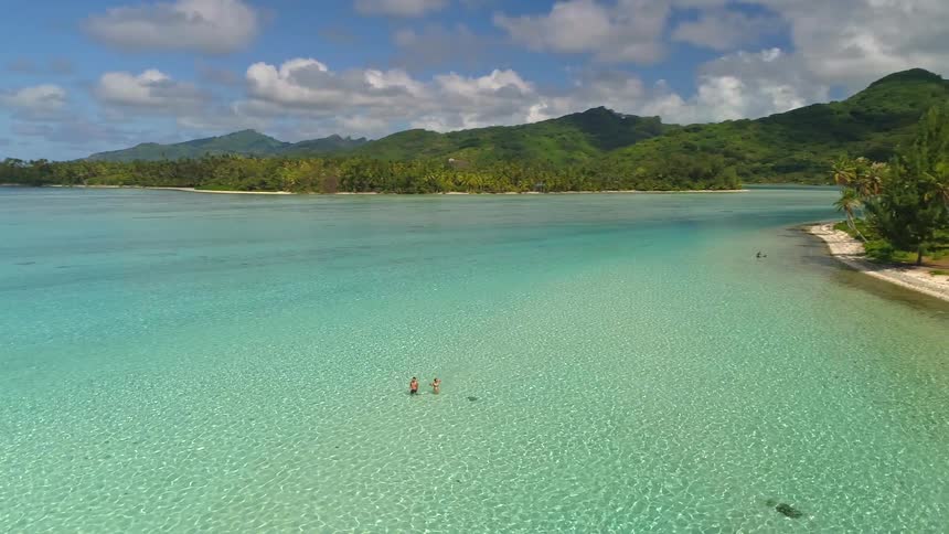 An aerial view pans over a couple and a stingray in the shallows of a stunningly clear turquoise lagoon, revealing a lush tropical island paradise