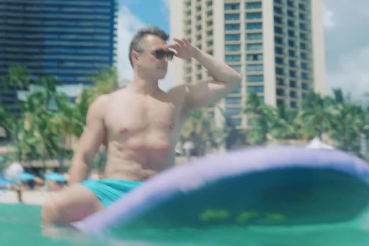 A shirtless man in sunglasses sits on his surfboard in the gentle ocean waves, looking out at a tropical city beach in slow motion