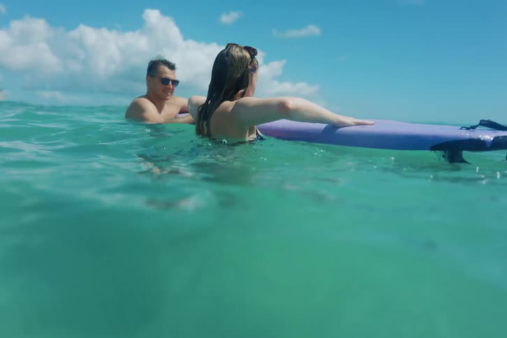 A slow-motion shot captures a couple enjoying a surfing lesson in the turquoise ocean waters of Hawaii with the Waikiki skyline in the background