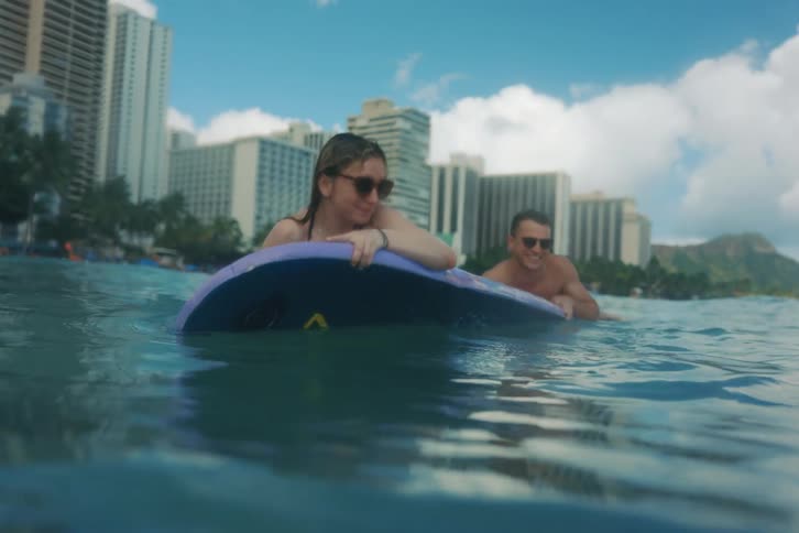 A couple in sunglasses relax on a shared surfboard, gently floating on the calm ocean waves off a tropical city beach