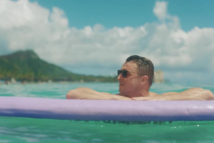 A tranquil shot of a man wearing sunglasses relaxing on a purple surfboard in the turquoise ocean on a beautiful sunny day