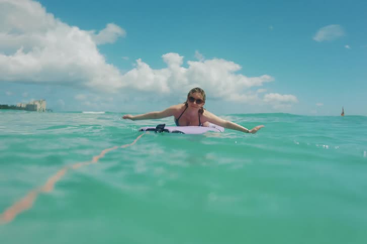 Young woman in a bikini happily learns to paddle a surfboard in the turquoise ocean waters of Waikiki Beach, Hawaii