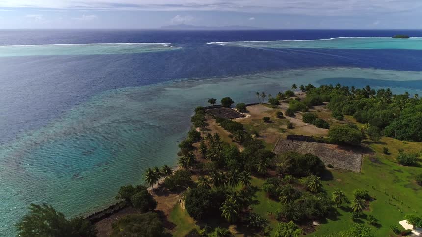 Aerial pullback shot over ancient marae ruins on a lush tropical island, revealing a turquoise lagoon and the deep blue ocean