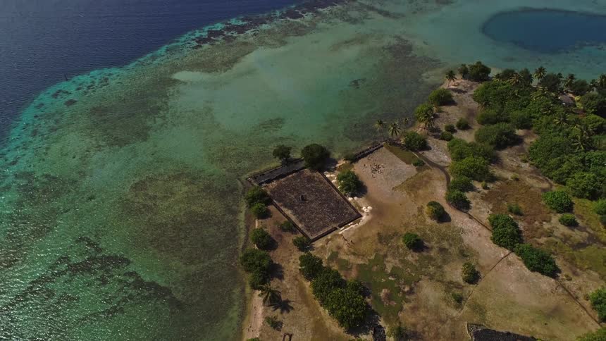 An aerial top-down view of a historic site on a tropical island slowly pans up to reveal the vast, beautiful ocean and coral reef