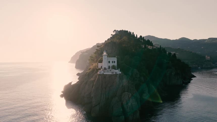 Aerial view pulls back from a picturesque white lighthouse on a rocky Italian coastline during a golden sunset