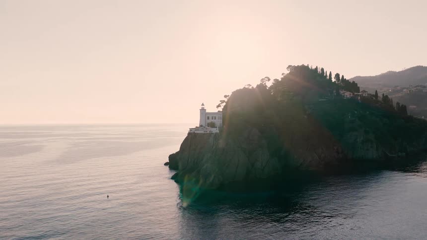 A serene aerial view of a white lighthouse perched on a rugged, tree-covered cliff over the calm ocean at sunrise