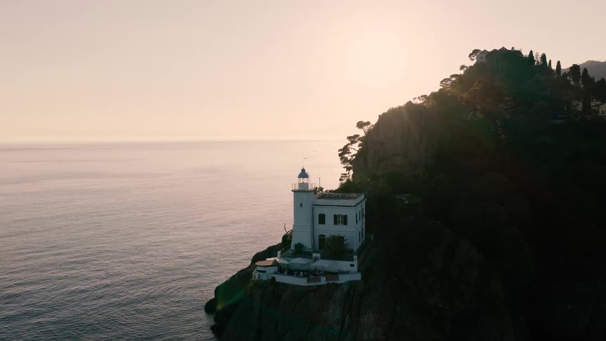 Slowly approaching the iconic Portofino Lighthouse with an aerial drone during a breathtaking sunrise over the calm Italian coastline
