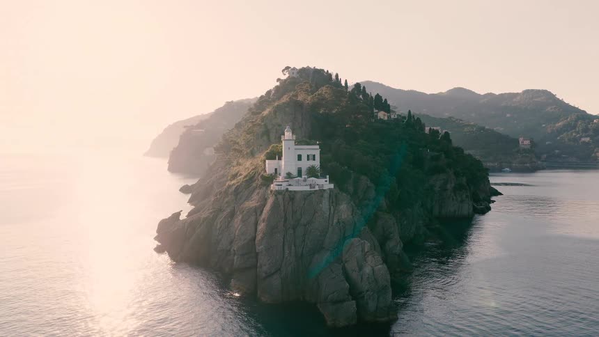 An aerial view of the Portofino lighthouse sitting atop a rocky cliff as the sun casts a golden glow over the sea
