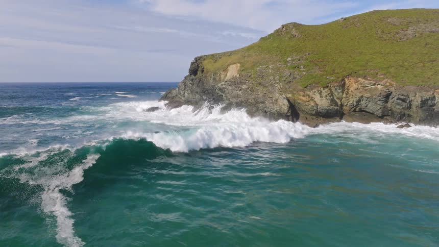 A massive ocean wave crashes against a rugged, grassy headland in stunning slow motion