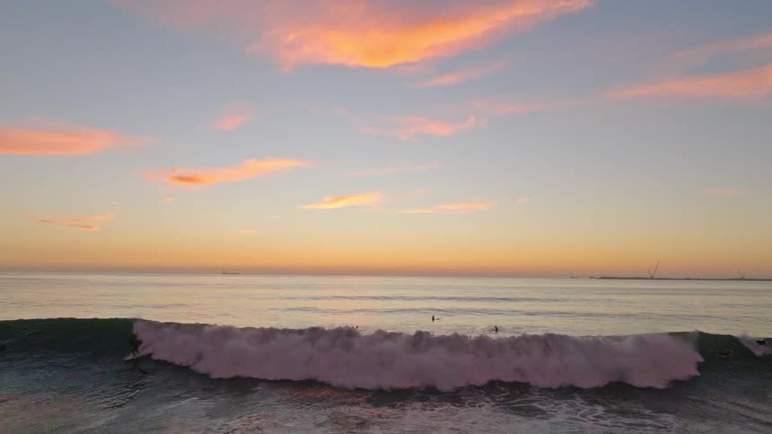 A group of surfers wait for the perfect wave as a large barrel rolls towards shore in slow motion during a beautiful sunset