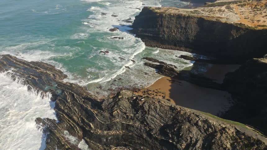 A stunning aerial shot slowly pans over a rugged coastline where powerful ocean waves crash against dramatic cliffs and layered rock formations