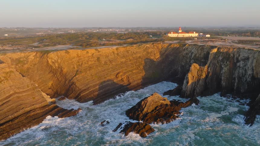The majestic Cabo do Sardão lighthouse stands atop dramatic, golden cliffs as the powerful Atlantic Ocean crashes against the rugged coastline at sunrise