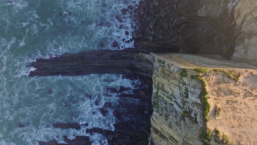A stunning top-down aerial view slowly rises above a rugged cliff as powerful ocean waves crash against the rocky coastline