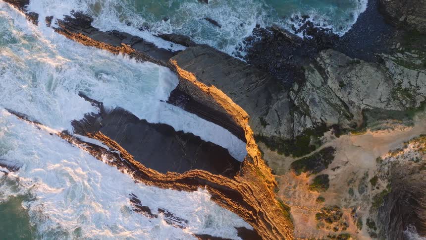 Stunning top-down drone footage ascends over a dramatic and rugged coastline, where waves crash against layered cliffs illuminated by the golden hour sun