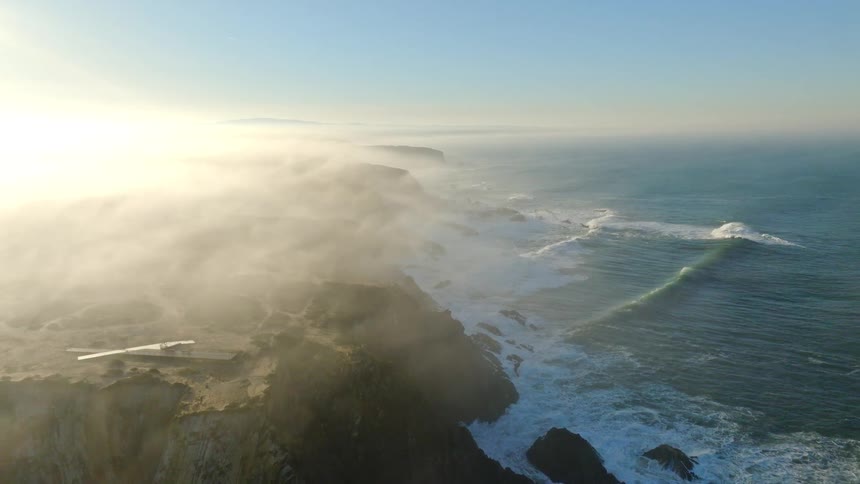 A majestic aerial view reveals a foggy, rocky coastline with powerful ocean waves rolling in at sunrise