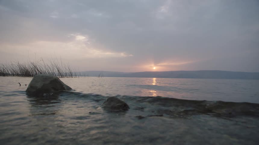 Gentle waves lap in slow motion against a rocky shore during a serene sunrise over a calm lake