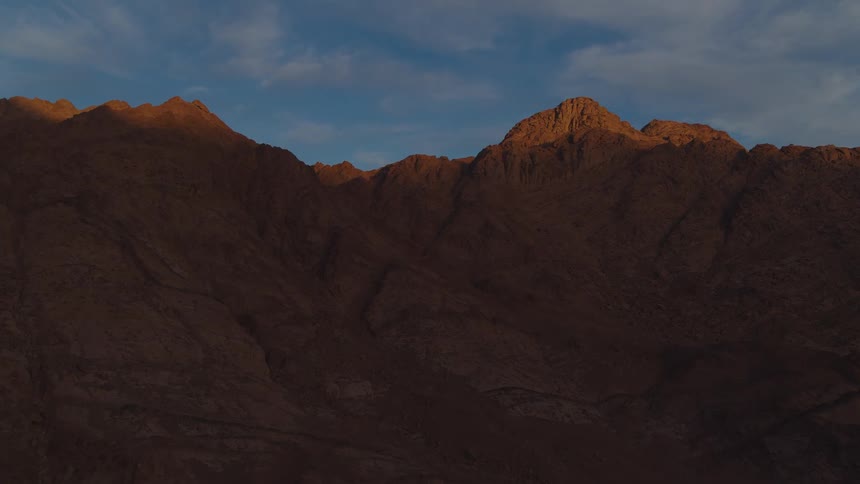 A slow aerial shot pushing towards a vast, rocky desert mountain range as the golden light of sunrise illuminates the peaks