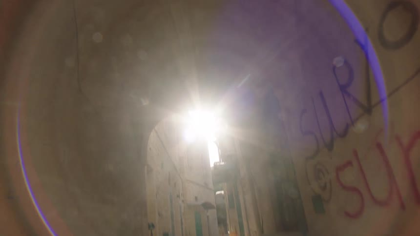 A low-angle dolly shot travels backward through a narrow stone alleyway as the camera slowly tilts up towards the sun
