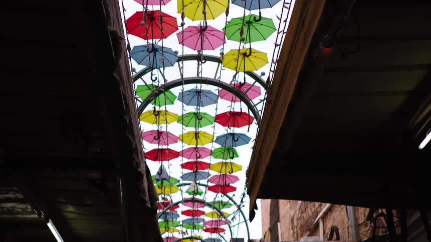 A low-angle, slow pull-back reveals a beautiful art installation of colorful umbrellas hanging over a narrow market street