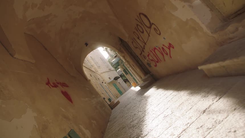 A slow, tilted camera movement pushes through a graffiti-covered stone alleyway toward a bright, sunlit opening