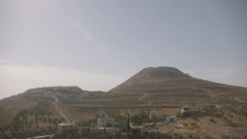 A slow tilt down from a hazy sun reveals the dry, terraced hills and a small village in the West Bank