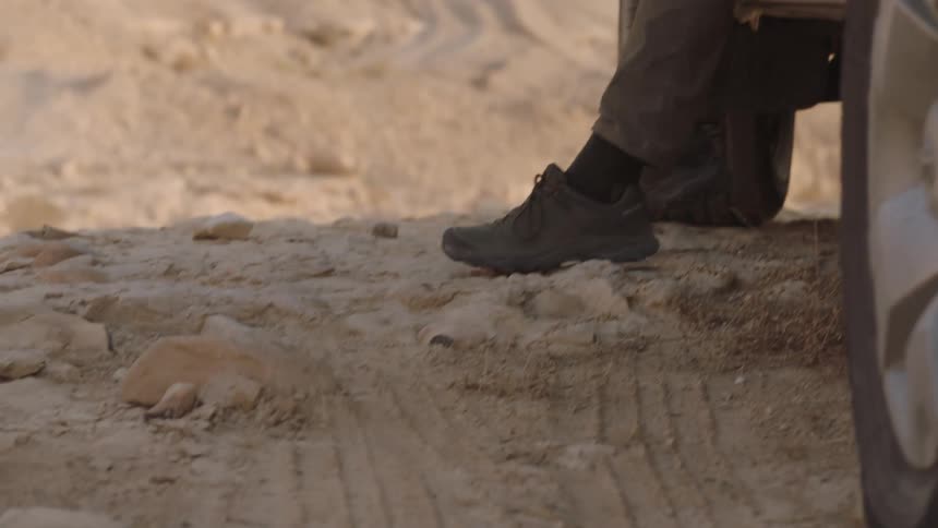 A man in hiking boots gets out of his pickup truck and walks across the rocky desert terrain in slow motion