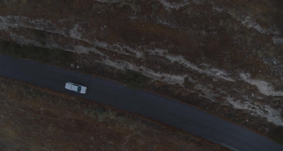 Top-down aerial shot follows a lone white car driving on a remote road through a dark, desolate landscape