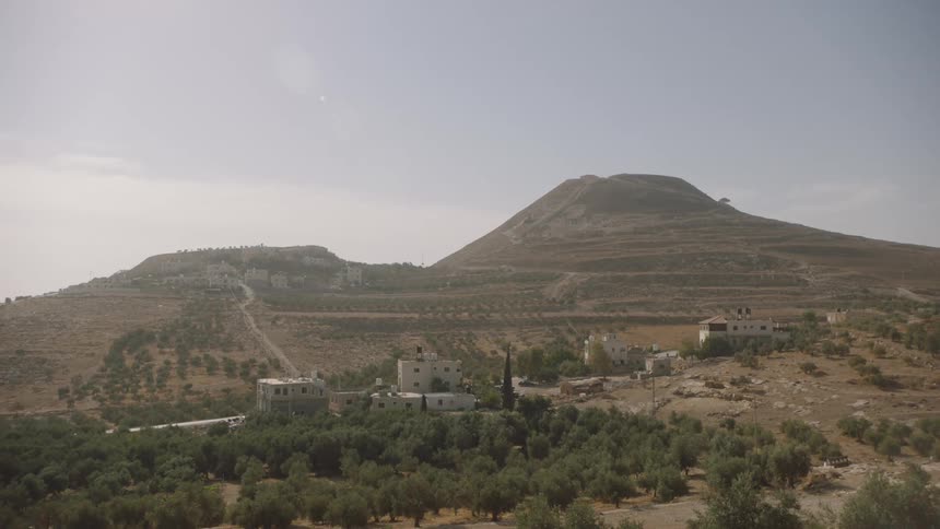 A wide, static view of a rural Middle Eastern village surrounded by olive groves at the base of a large, terraced hill