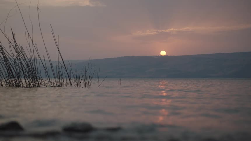 The sun sets over distant hills, casting a warm glow over a tranquil lake as gentle waves lap the shore