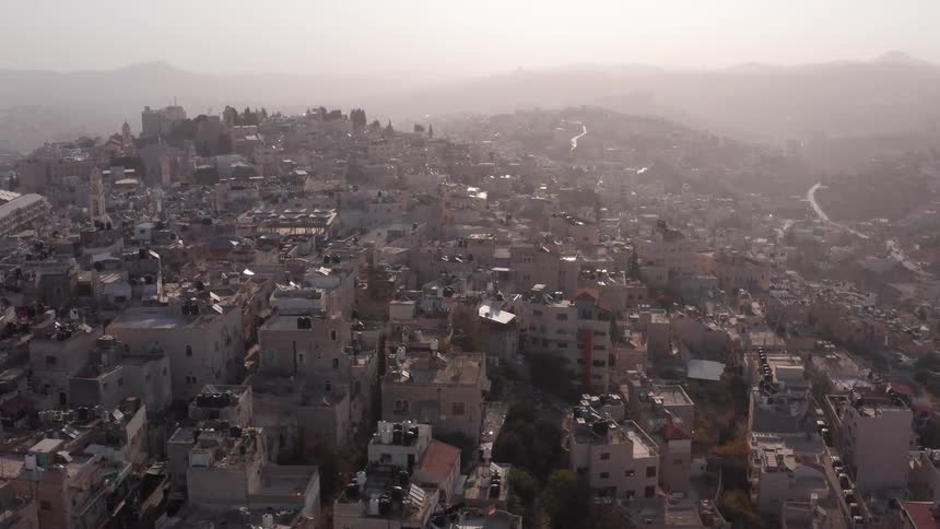 An aerial view reveals a dense, historic Middle Eastern city built on hillsides during a hazy sunrise