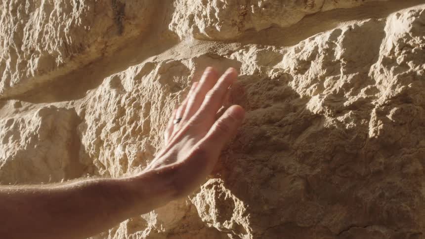 A man's hand gently touches and slides across an ancient, sunlit stone wall in slow motion