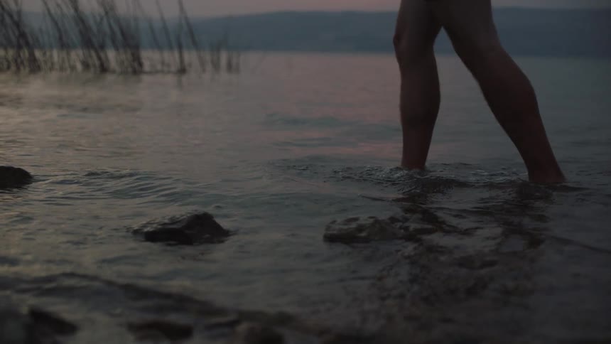 A slow-motion close-up of bare feet and legs walking through shallow water at dusk, splashing gently with each step