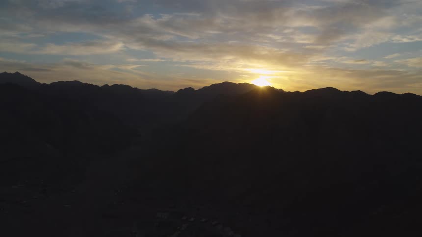 A static aerial view shows the sun rising over a silhouetted mountain range, casting a golden glow on the morning sky