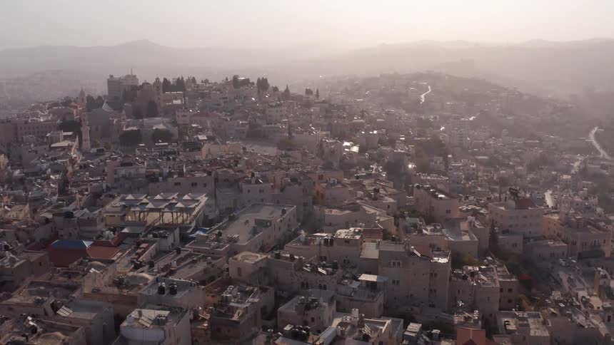 A slow-pullback drone shot reveals the ancient city of Bethlehem, with its historic buildings and rooftops stretching across the hills under a hazy morning sky