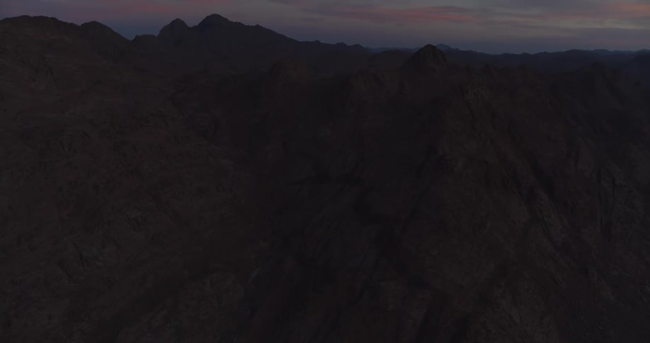 A slow aerial tilt-up shot reveals a vast, rocky mountain range silhouetted against a colorful twilight sky