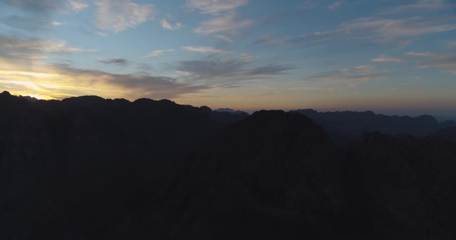 A slow aerial ascent reveals a vast, dark mountain range silhouetted against the beautiful colors of a sunrise