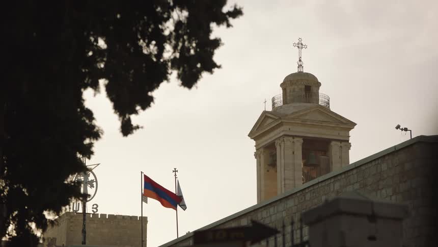 The Armenian flag waves gently next to a historic stone church bell tower under a bright, overcast sky in Jerusalem