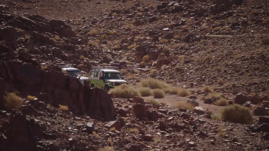 A pair of off-road safari jeeps are parked on a dirt path winding through a sun-drenched, rocky desert canyon