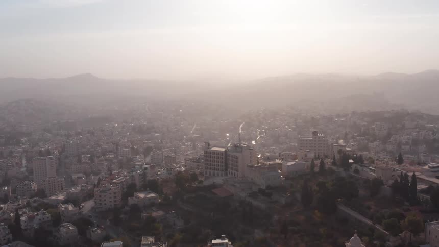 A slow pull-back aerial view reveals a sprawling Middle Eastern city shrouded in morning haze with mountains visible in the distance