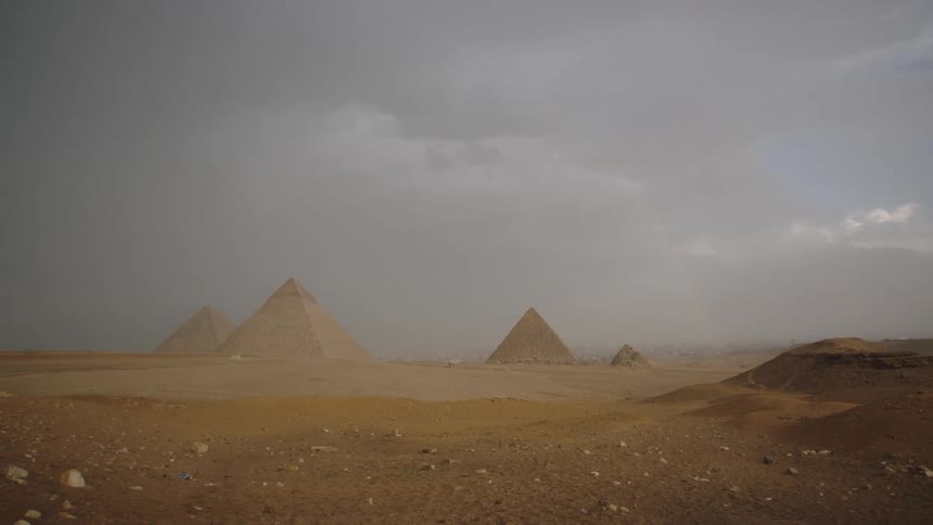 The majestic Great Pyramids of Giza stand tall in the desert under a dramatic, stormy sky with a faint rainbow