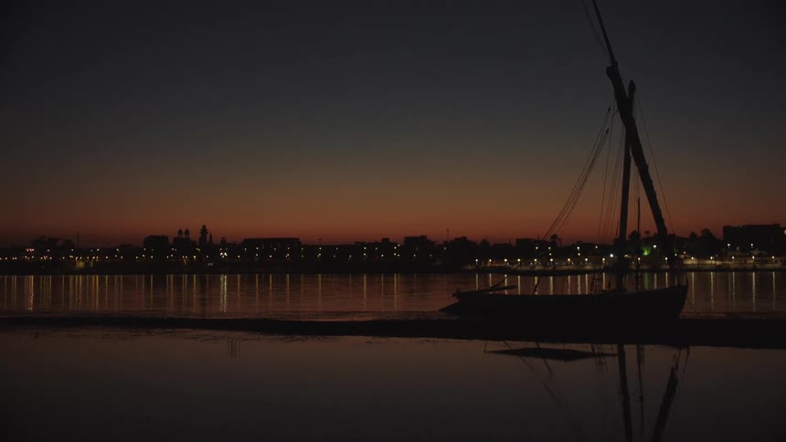 A traditional Egyptian felucca boat is silhouetted against the colorful dusk sky on the Nile River, with the city lights of Luxor reflecting in the calm water
