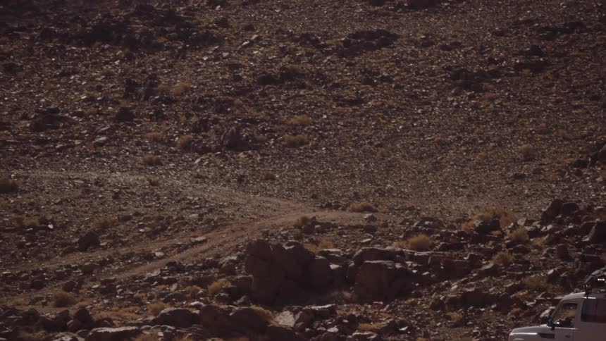 A 4x4 vehicle drives up a steep, rocky mountain road in a barren landscape
