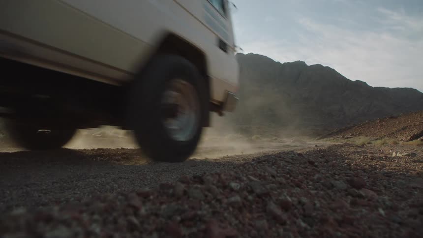 Slow motion, low-angle shot of an off-road vehicle driving on a dusty desert road with mountains in the background as the sun sets