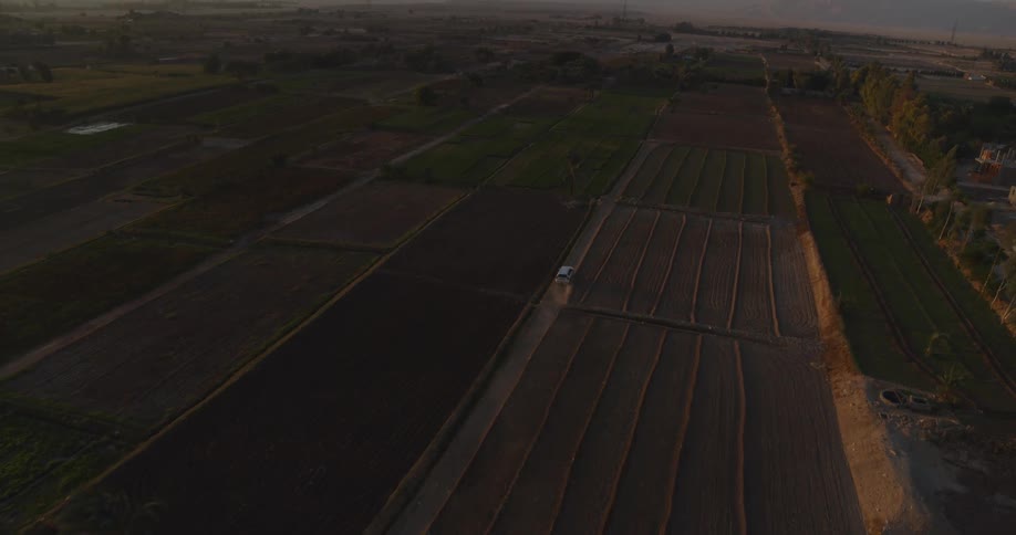 Aerial view of a white van driving along a dirt road through expansive rural farmlands at sunset
