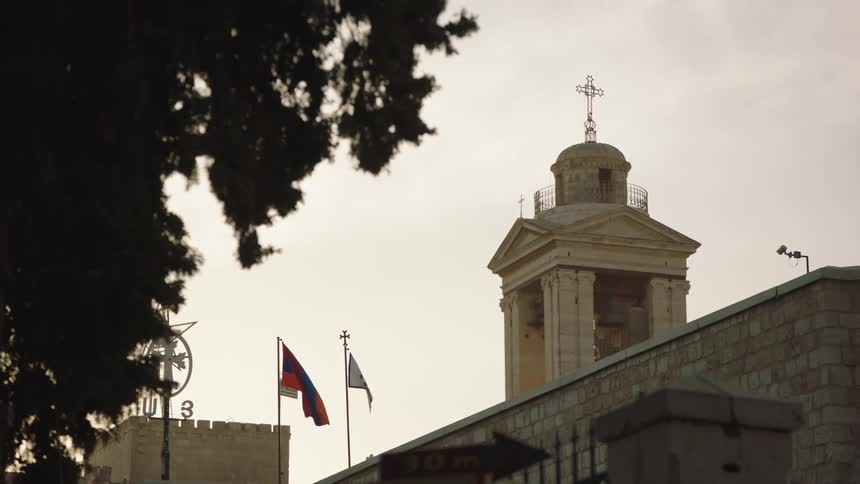 A historic church bell tower with a cross stands tall as an Armenian flag waves gently in the wind against a pale sky